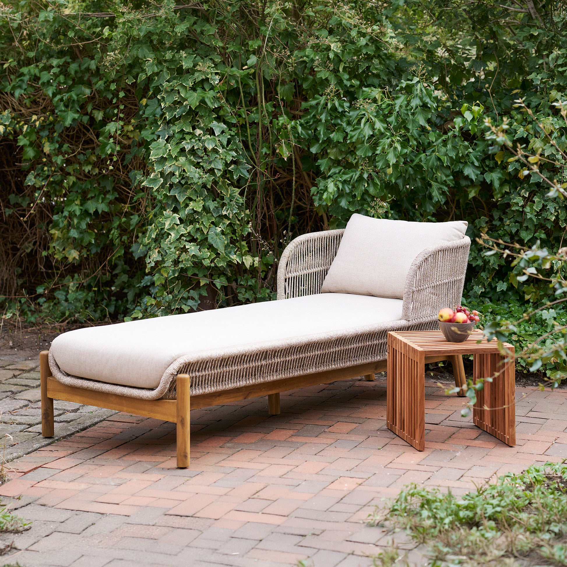 Outdoor lounge chair with cushions on a patio next to a small table with fruit, set against a green hedge.