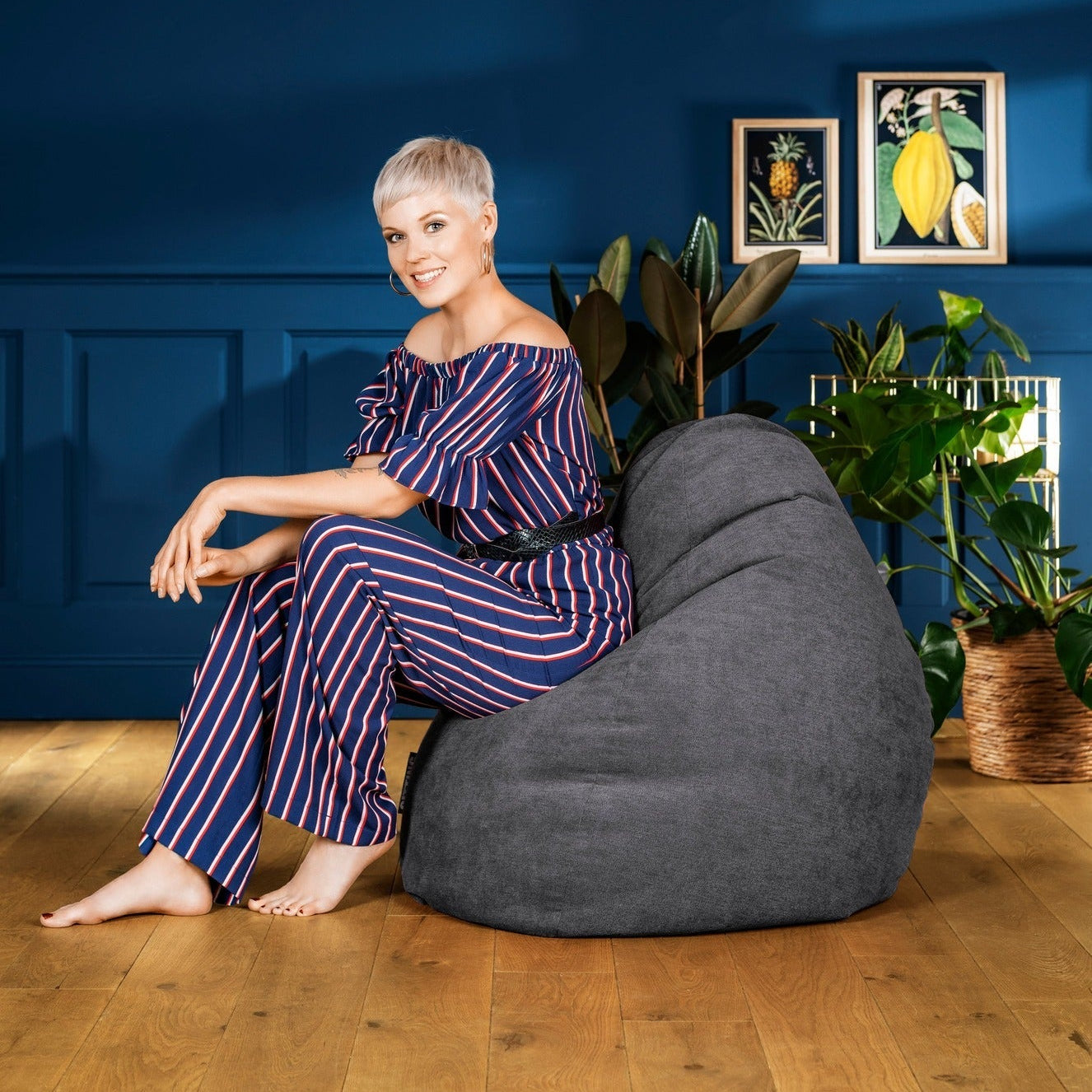 Woman sitting on a gray bean bag chair against a blue wall with plants and framed pictures.