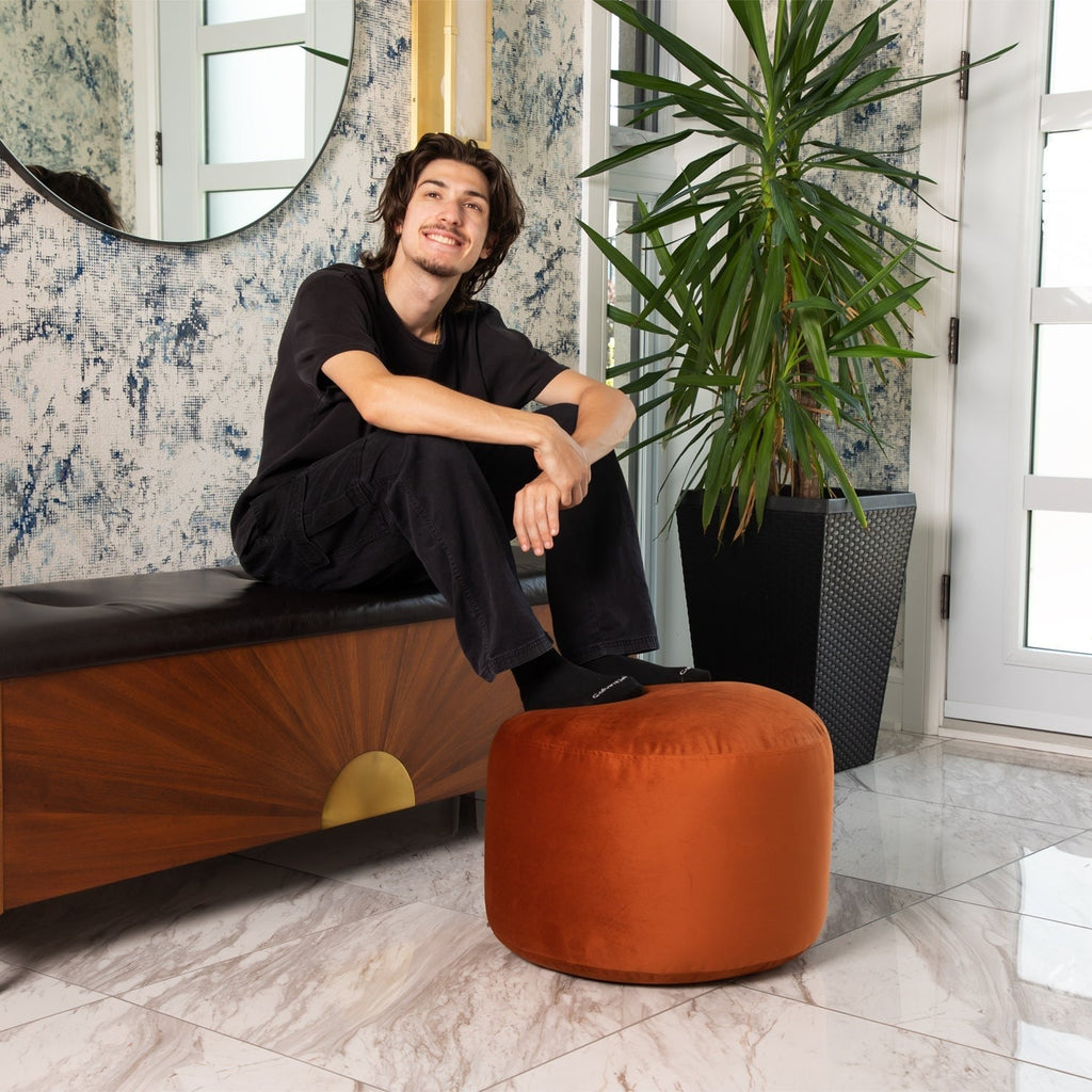 Person sitting on a wooden bench with an orange pouf and a plant in a stylish interior setting.