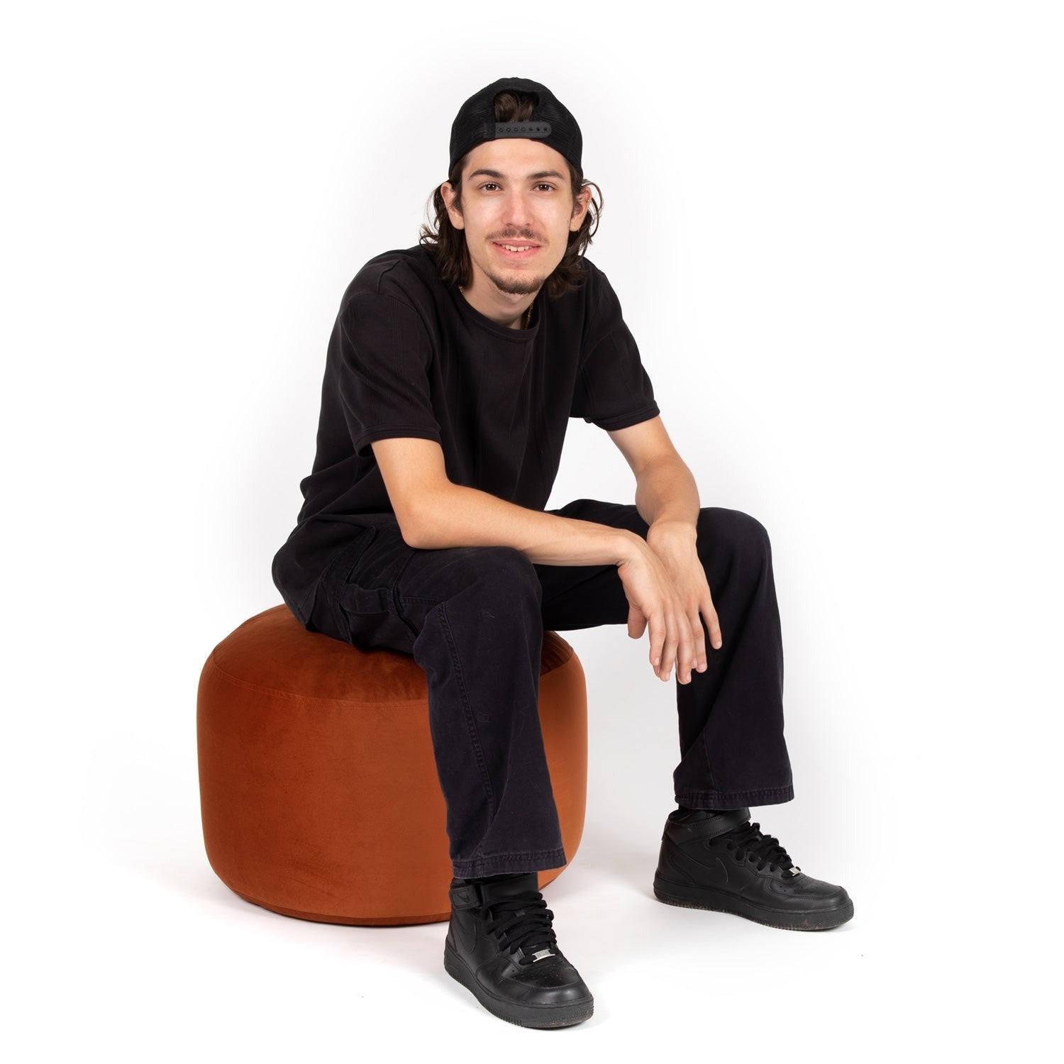 Man sitting on a brown stool wearing black clothing against a white background