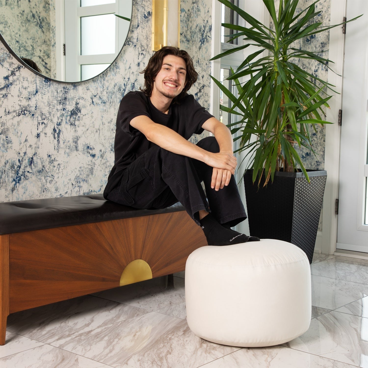 Man sitting on a bench in a stylish room with decorative wallpaper and a plant.