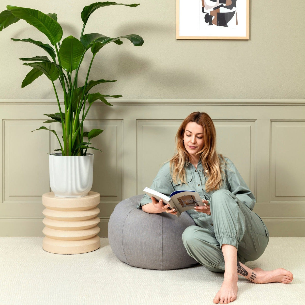 Woman reading a book on a gray pouf in a room with a plant and framed picture on the wall.