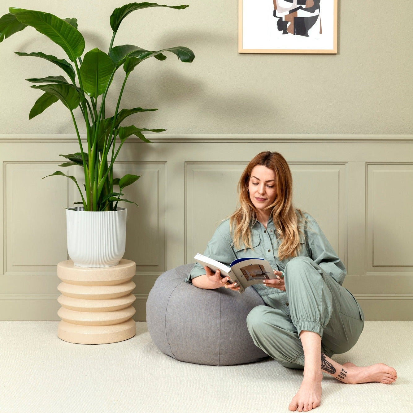 Woman reading a book on a gray pouf in a room with a plant and framed picture on the wall.