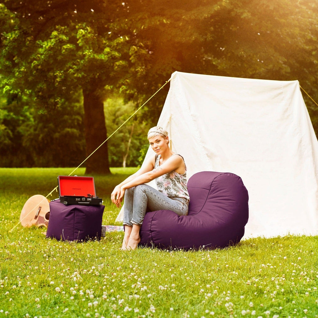 Person sitting on a bean bag chair in a grassy area with a tent and open laptop.