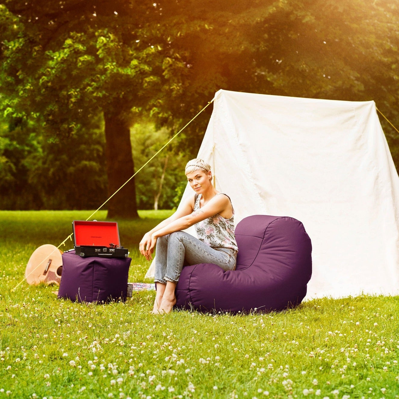 Person sitting on a bean bag chair in a grassy area with a tent and open laptop.
