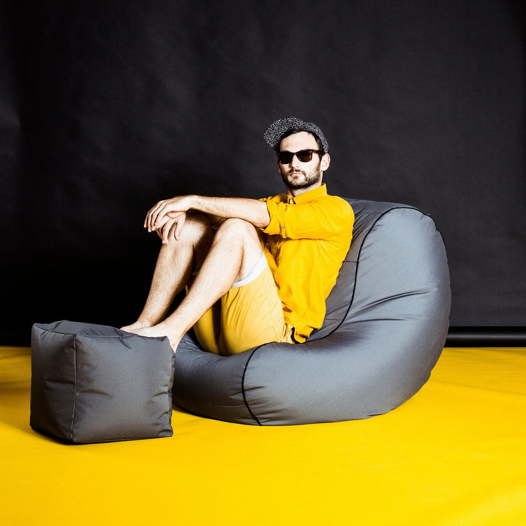 Man in yellow shirt and shorts sitting on a gray bean bag chair with a black pillow on a yellow floor and black background