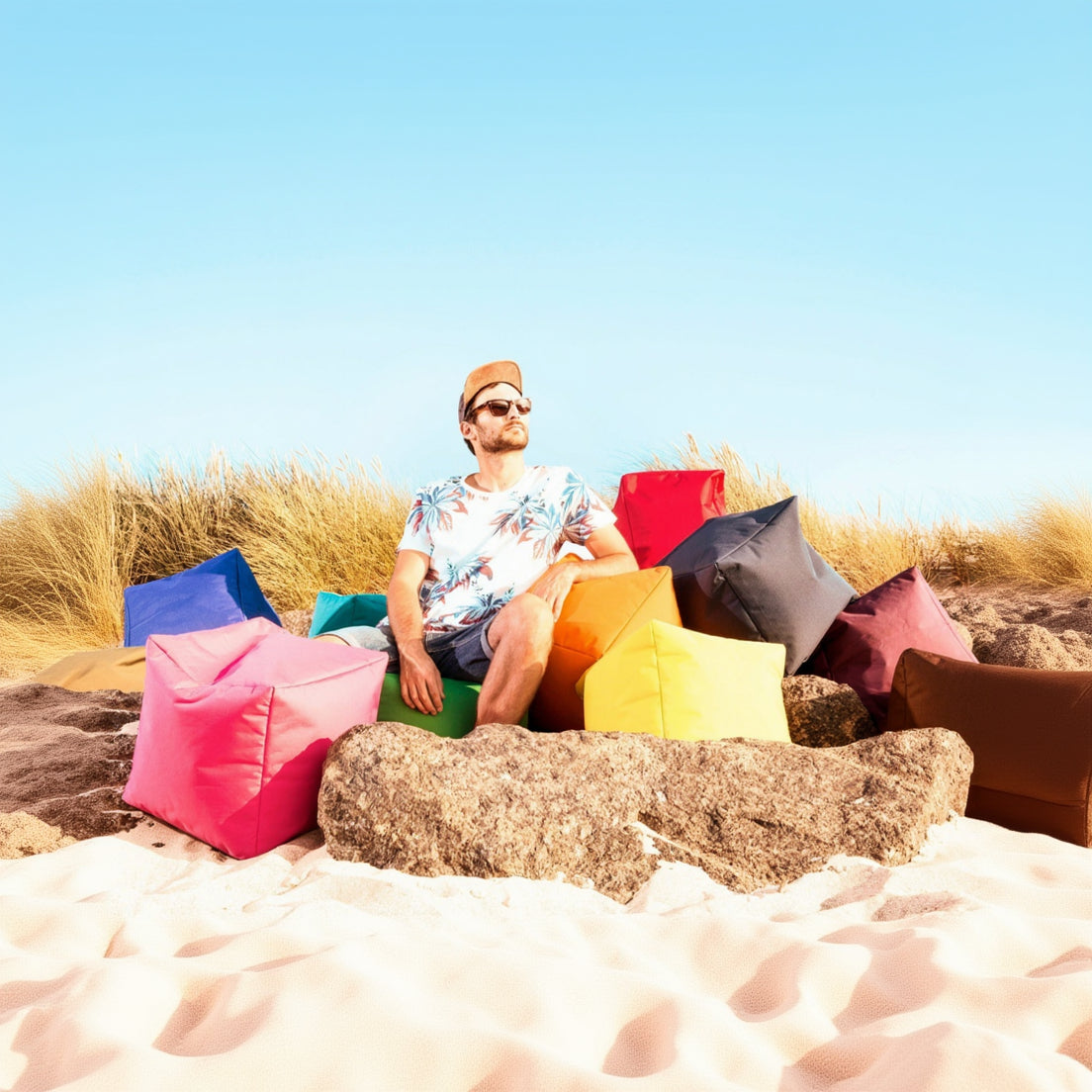 Man sitting on a colorful bean bag chair in a sandy area with a clear blue sky.
