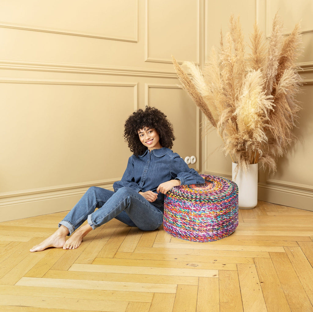 Woman sitting on a colorful ottoman in a room with a vase of dried plants.