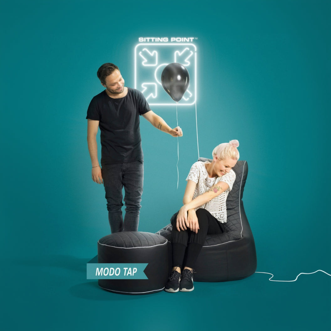 Two people interacting with a black bean bag chair and ottoman against a teal background, featuring 'Sitting Point' branding.