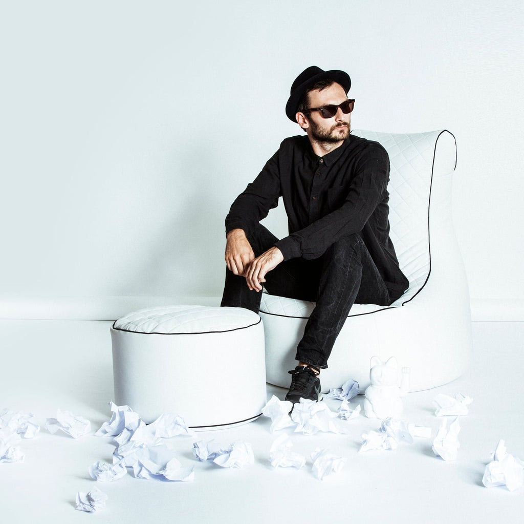 Man in black outfit sitting on a white chair with scattered white balls around, against a white background