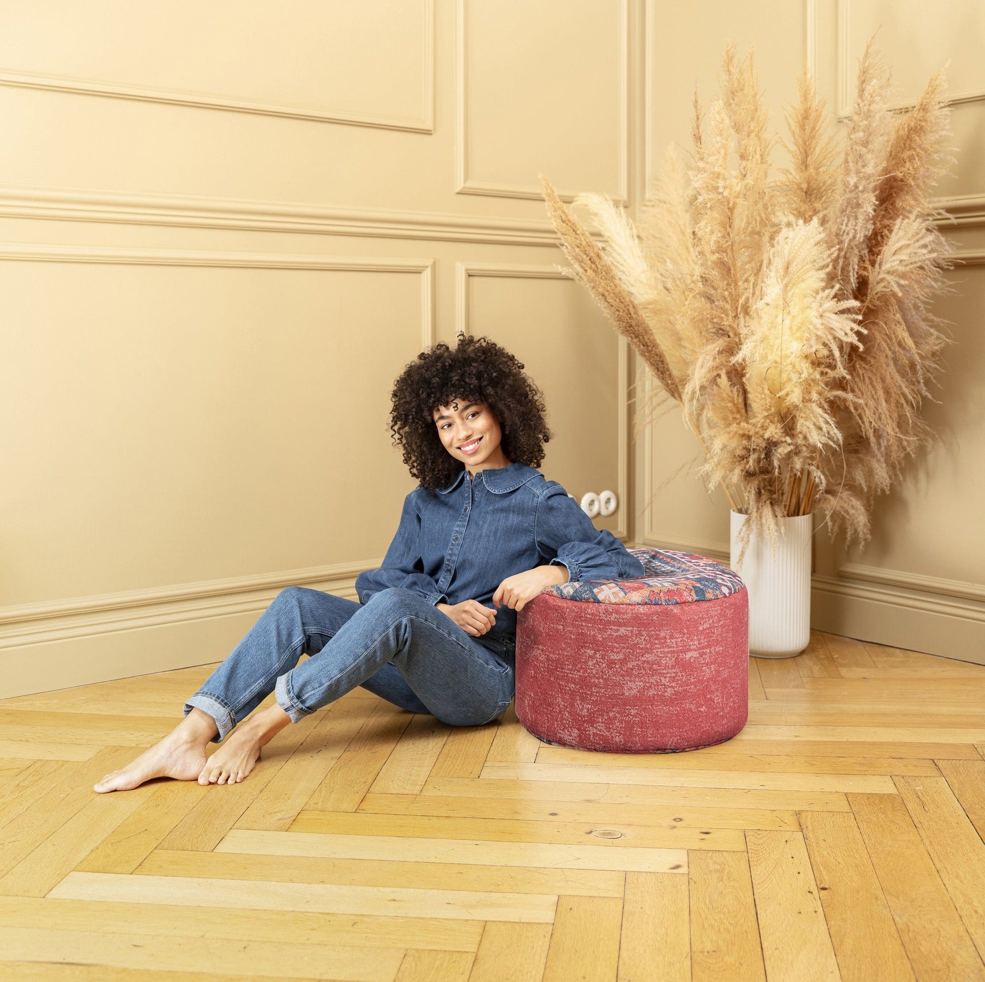 Woman sitting on a colorful ottoman in a room with beige walls and wooden floor.