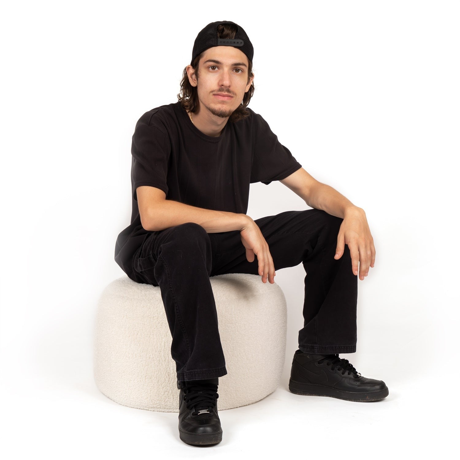 Person wearing a black outfit sitting on a white stool against a white background