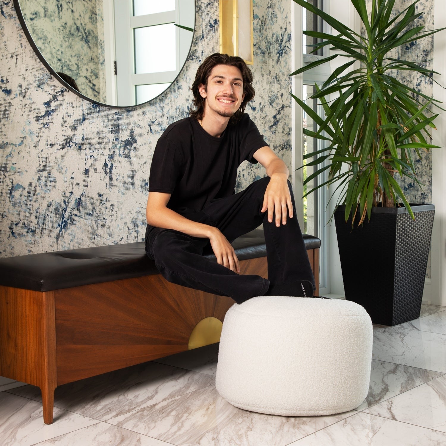 Man sitting on a wooden bench in a stylish interior setting with a plant and mirror.