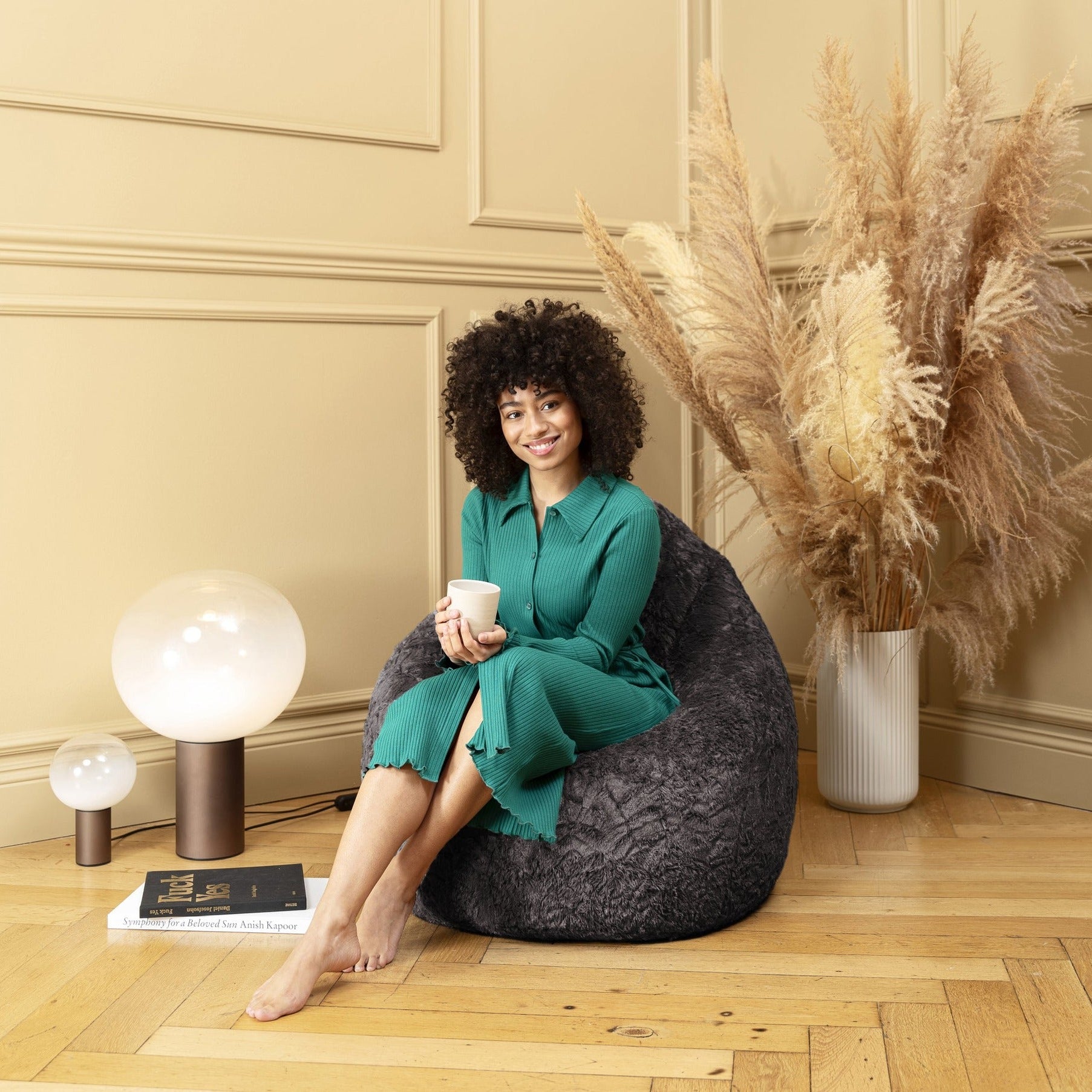 Woman sitting on a bean bag chair holding a cup, surrounded by decorative elements in a room with beige walls and wooden floor.