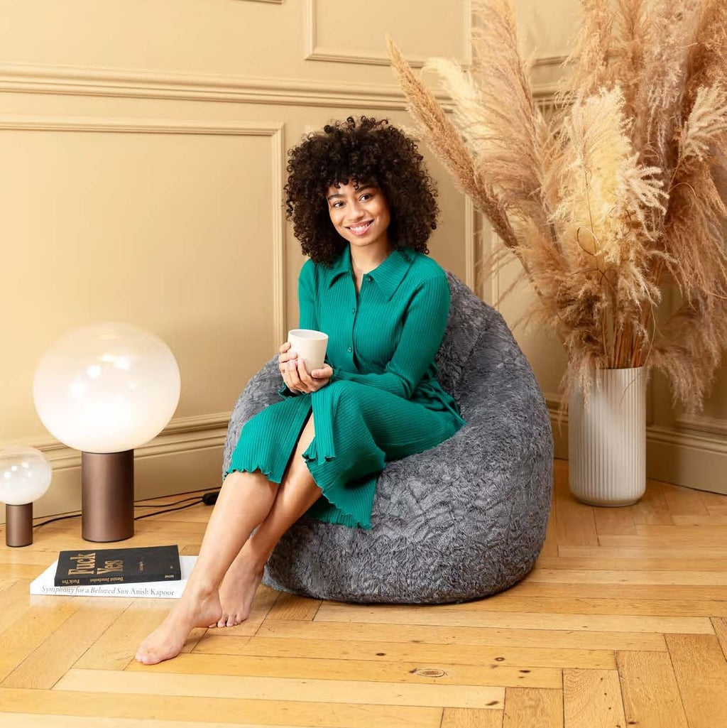 Woman in a green dress sitting on a gray bean bag chair holding a cup, with decorative elements like pampas grass and a vase in the background.
