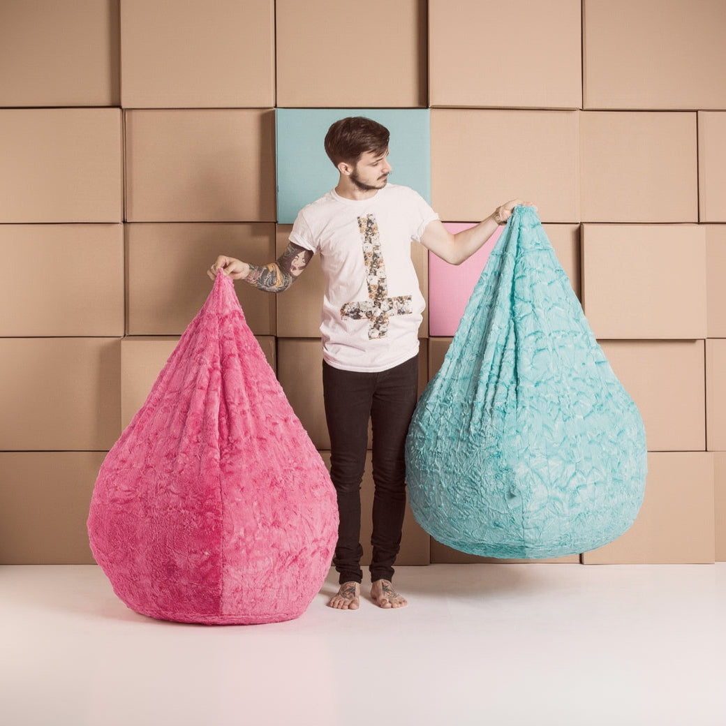 Man holding a pink and a blue bean bag chair against a wall of stacked boxes.