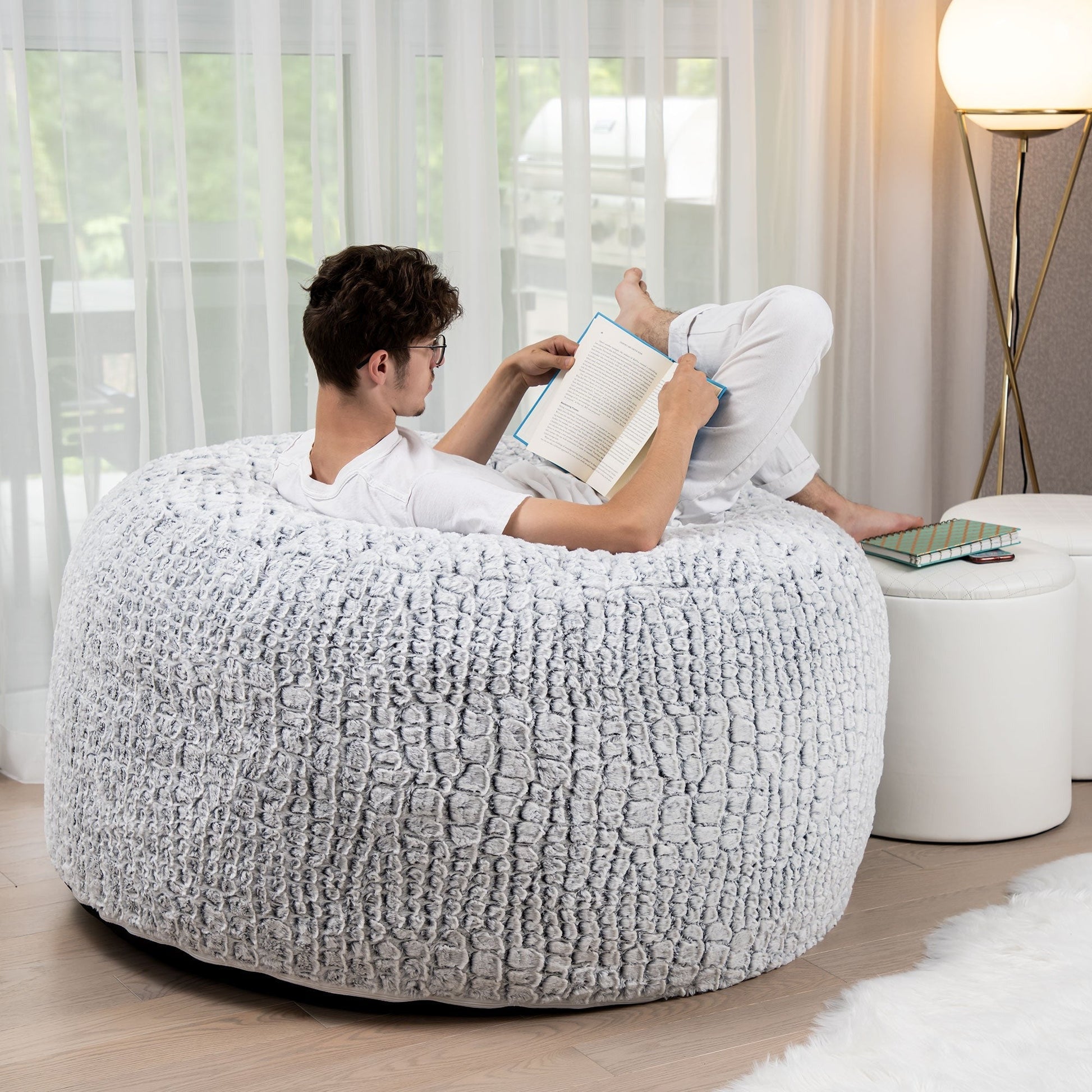 Person reading a book on a textured white bean bag chair in a modern living room.
