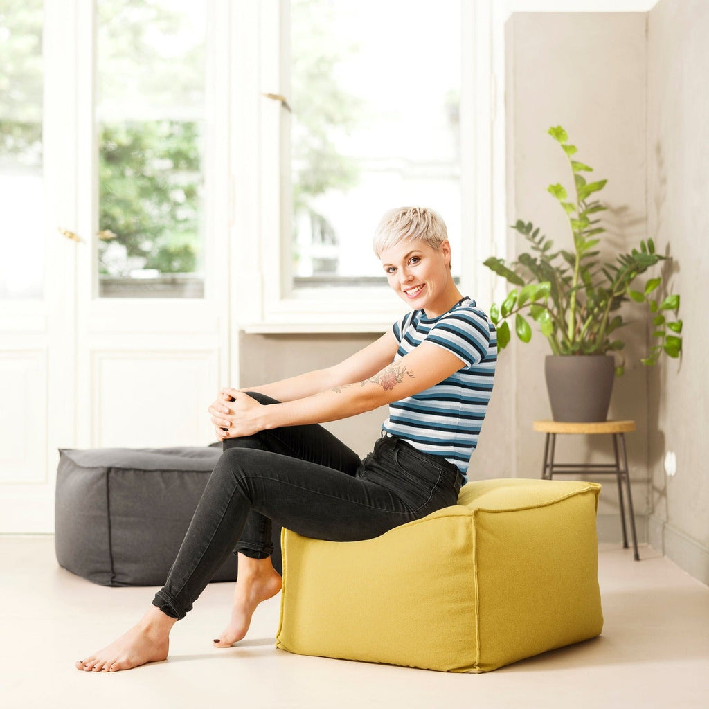 Person sitting on a yellow bean bag chair in a bright room with large windows and plants.