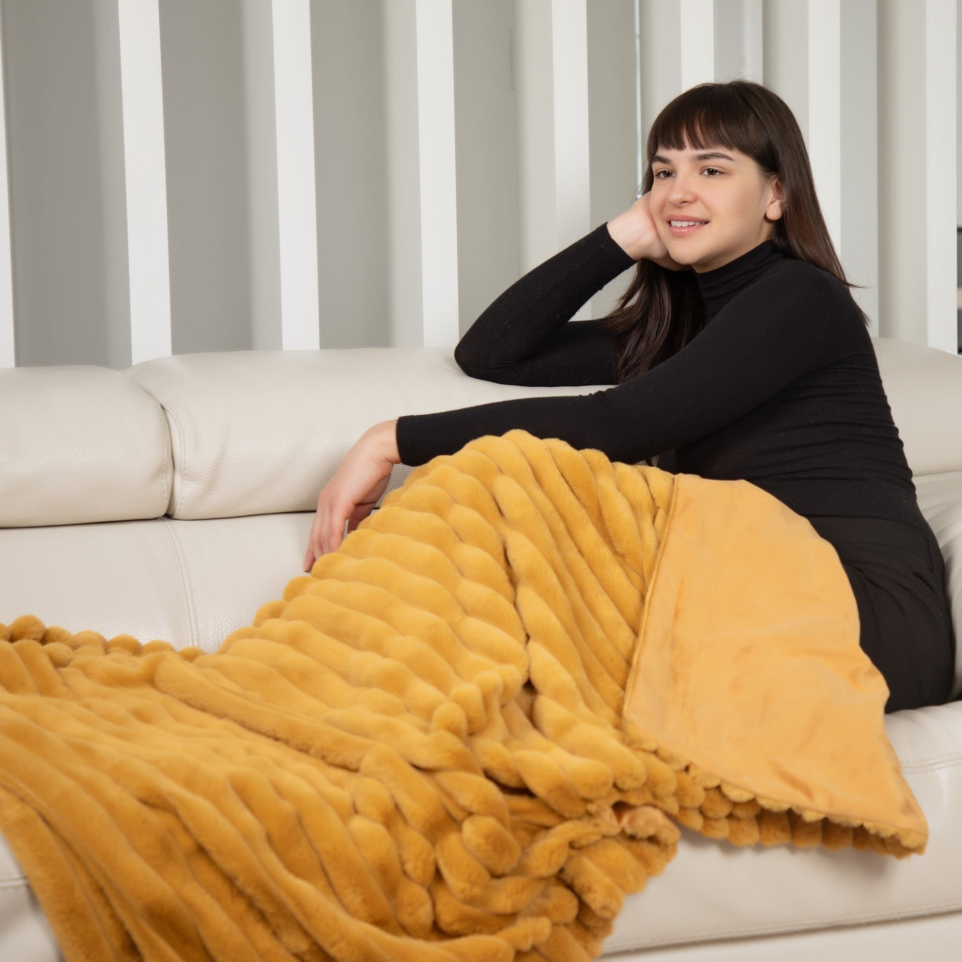Woman sitting on a couch with a yellow blanket draped over her, against a striped curtain background.