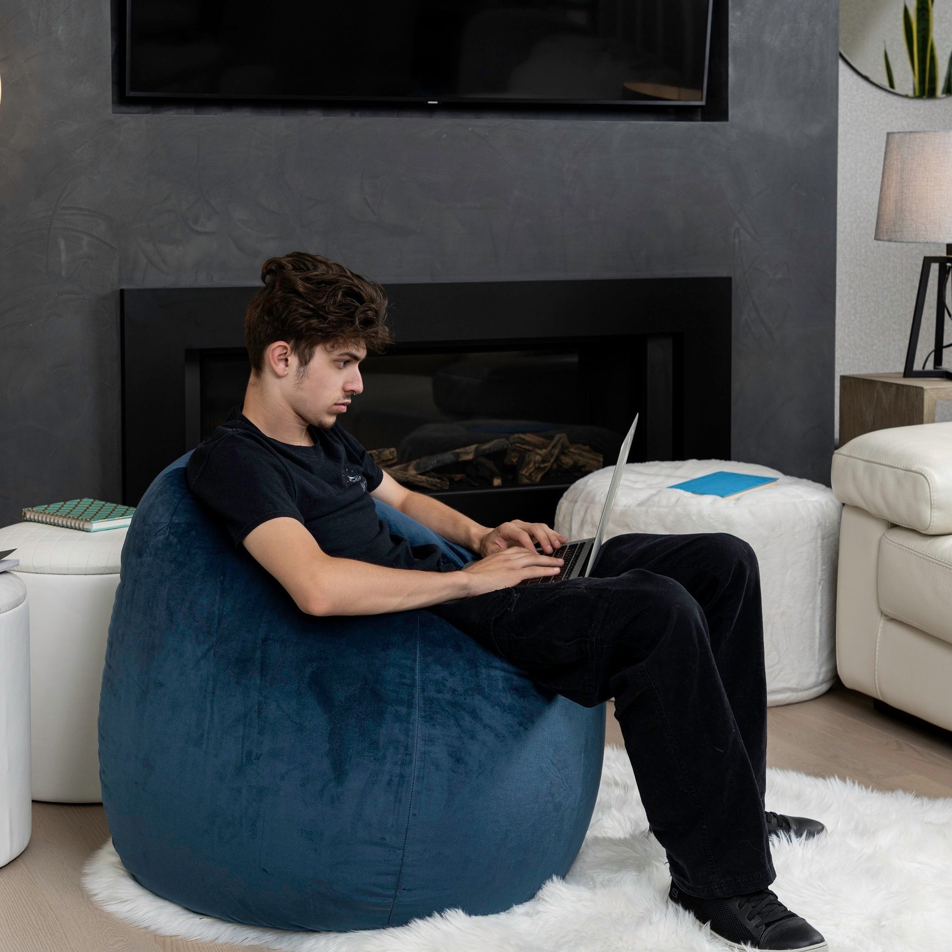 Man sitting on a blue bean bag chair using a laptop in a living room.