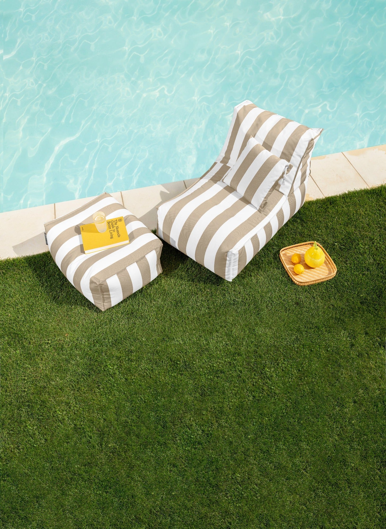Two striped lounge chairs by a poolside with snacks on a grassy area.