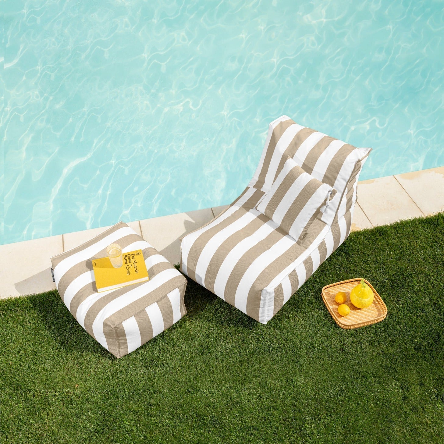 Two striped lounge chairs by a poolside with snacks on a grassy area.