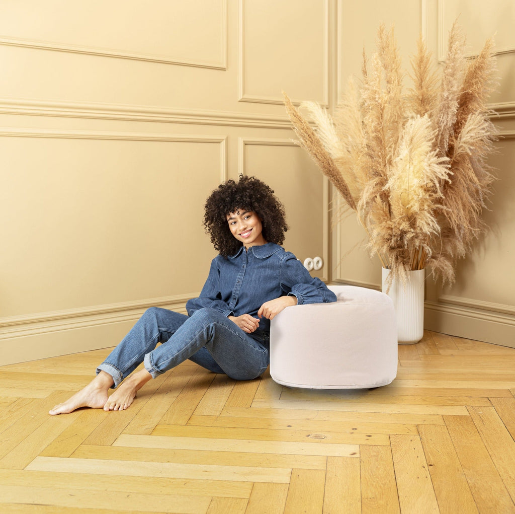Woman sitting on a white ottoman in a room with beige walls and wooden floor.
