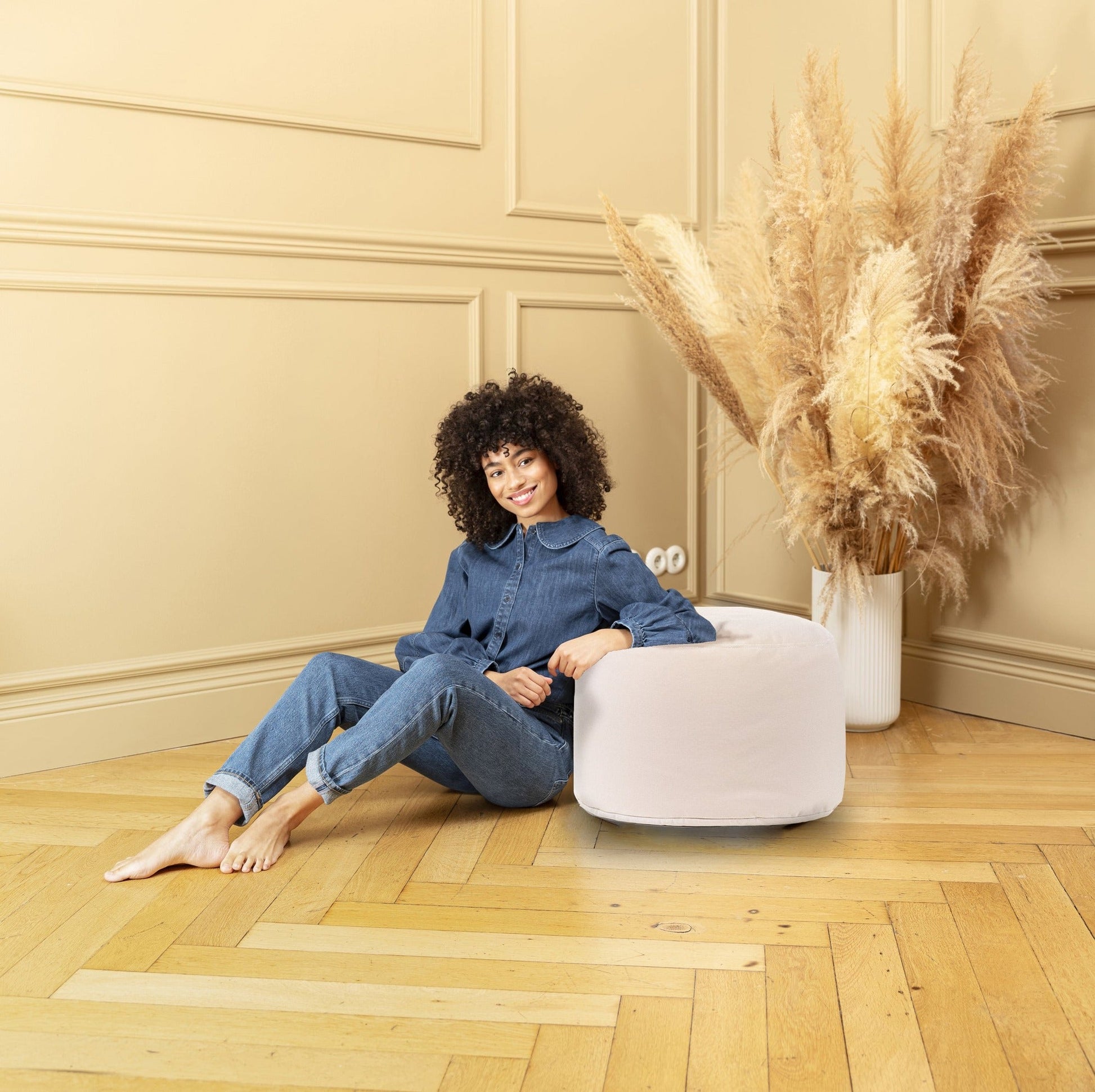 Woman sitting on a white ottoman in a room with beige walls and wooden floor.