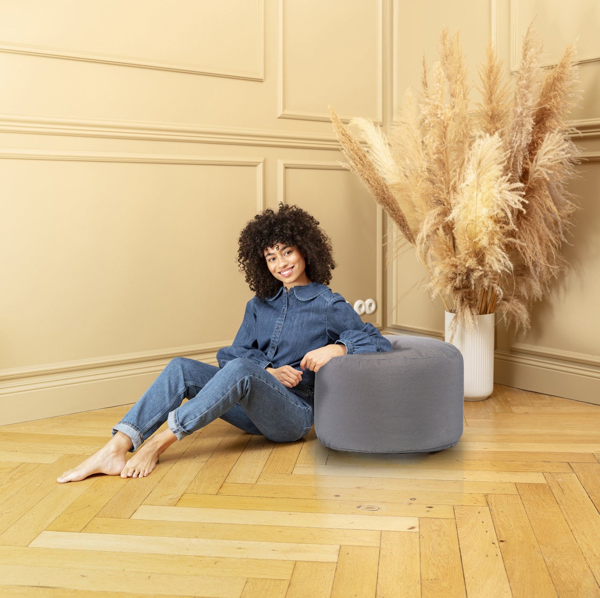 Woman sitting on a gray pouf in a room with beige walls and wooden floor.