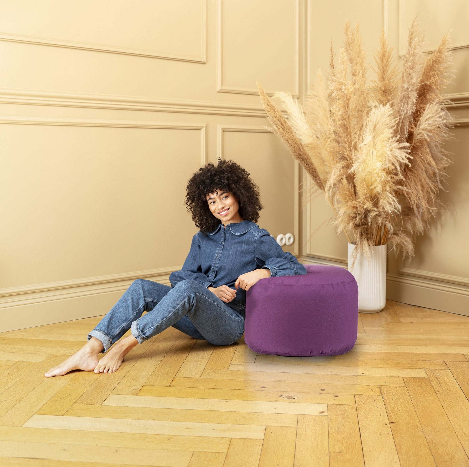 Woman sitting on a purple pouf in a room with beige walls and wooden floor.