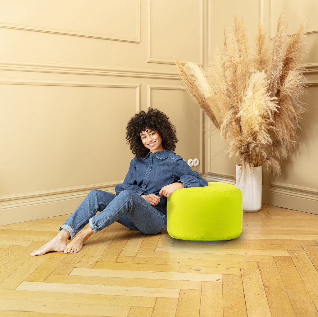 Woman sitting on a green ottoman in a room with beige walls and wooden floor.