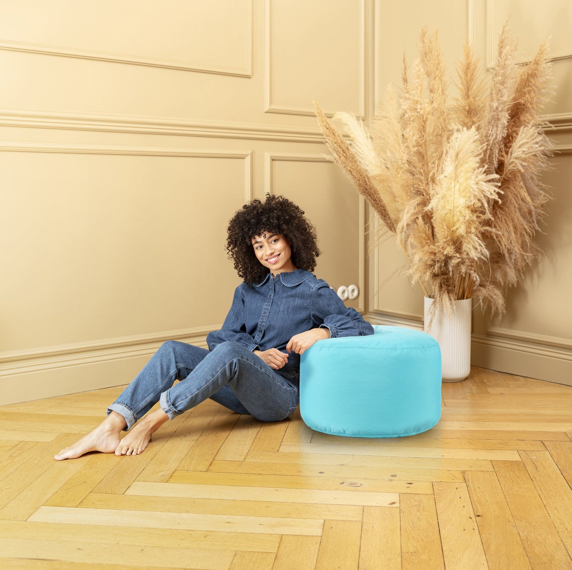 Woman sitting on a blue pouf in a room with beige walls and wooden floor.