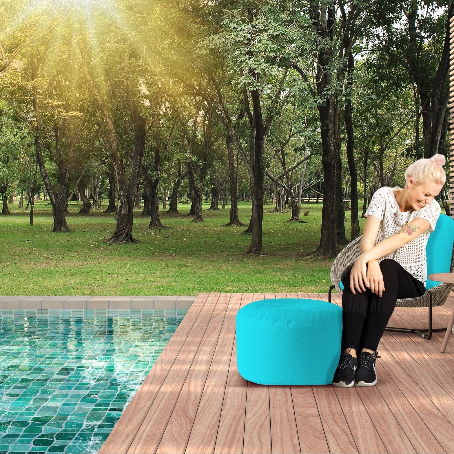Woman sitting by a pool with a blue ottoman and chair in a sunlit garden.