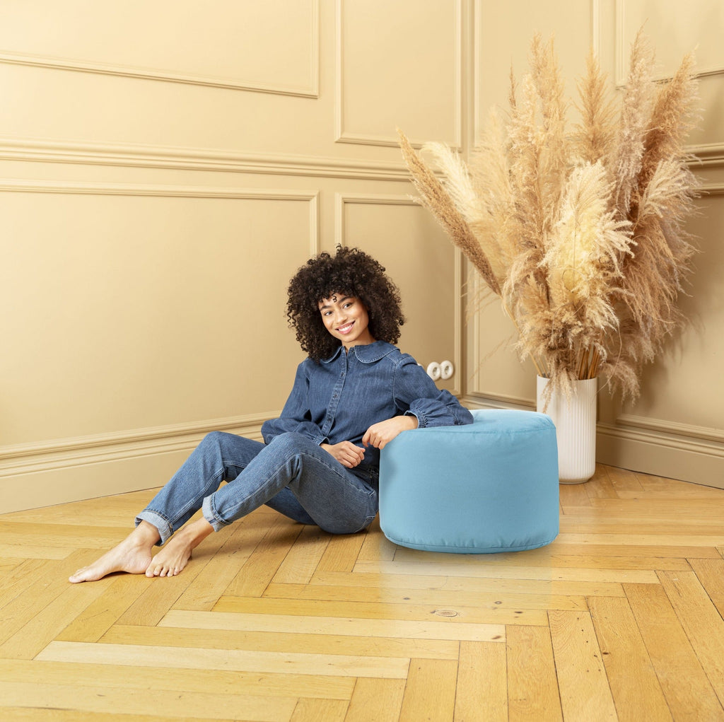 Woman sitting on a blue pouf in a room with beige walls and wooden floor.