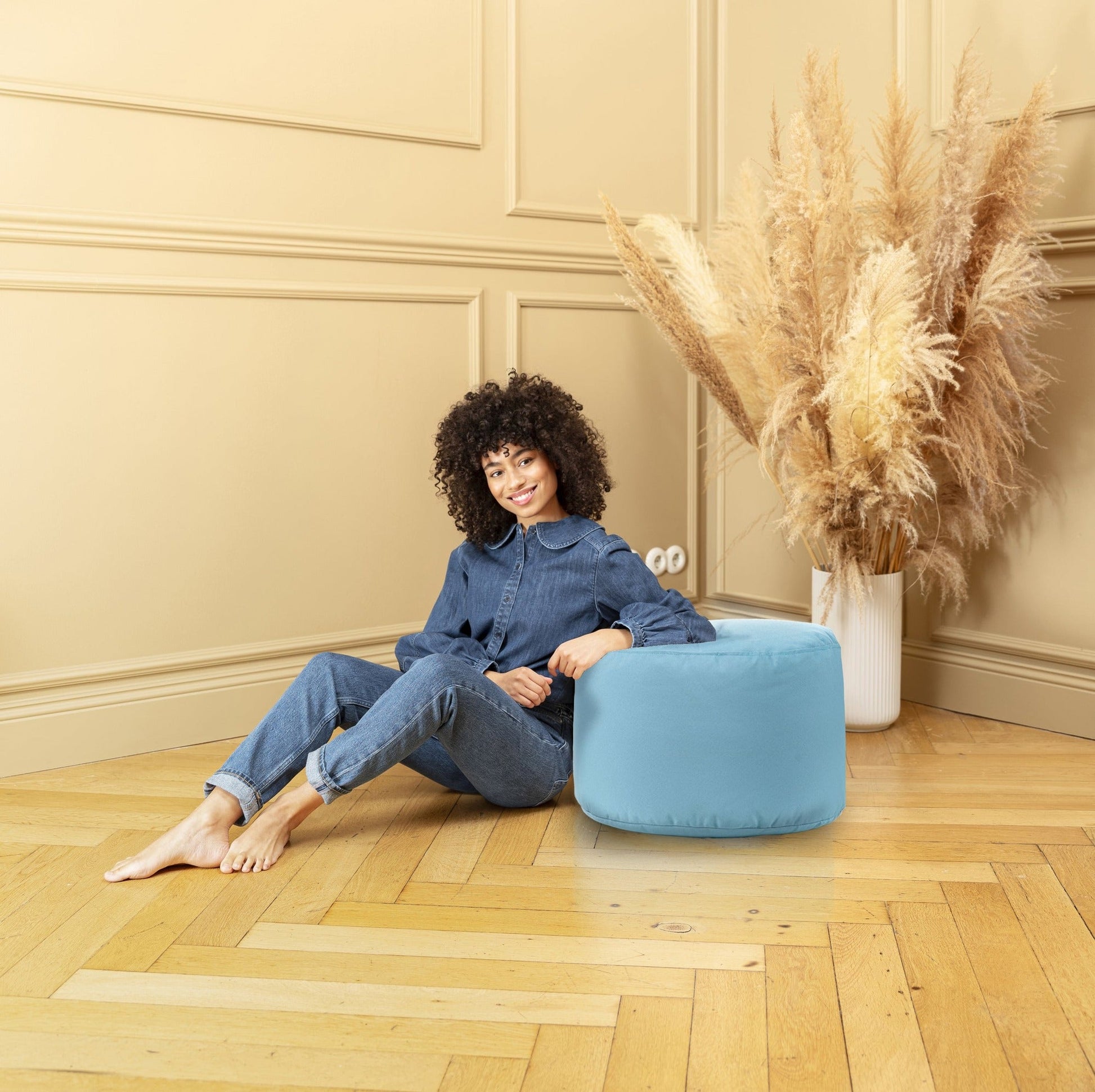 Woman sitting on a blue pouf in a room with beige walls and wooden floor.