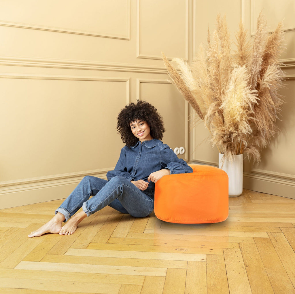 Woman sitting on an orange pouf in a room with beige walls and a vase of dried plants.