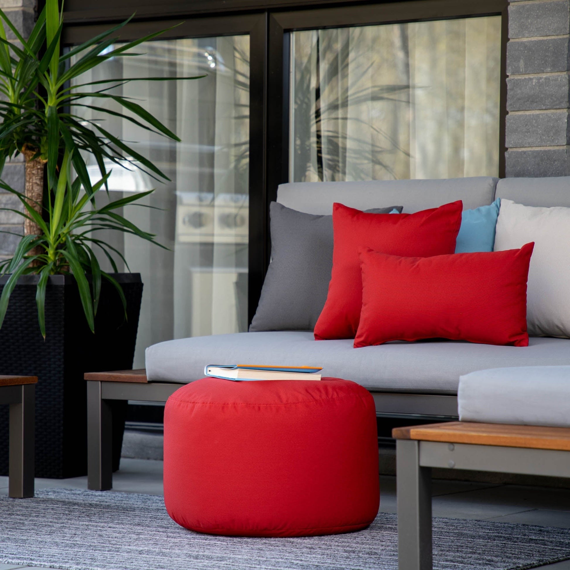 Outdoor seating area with a gray sofa, red pouf, and colorful cushions on a patio.