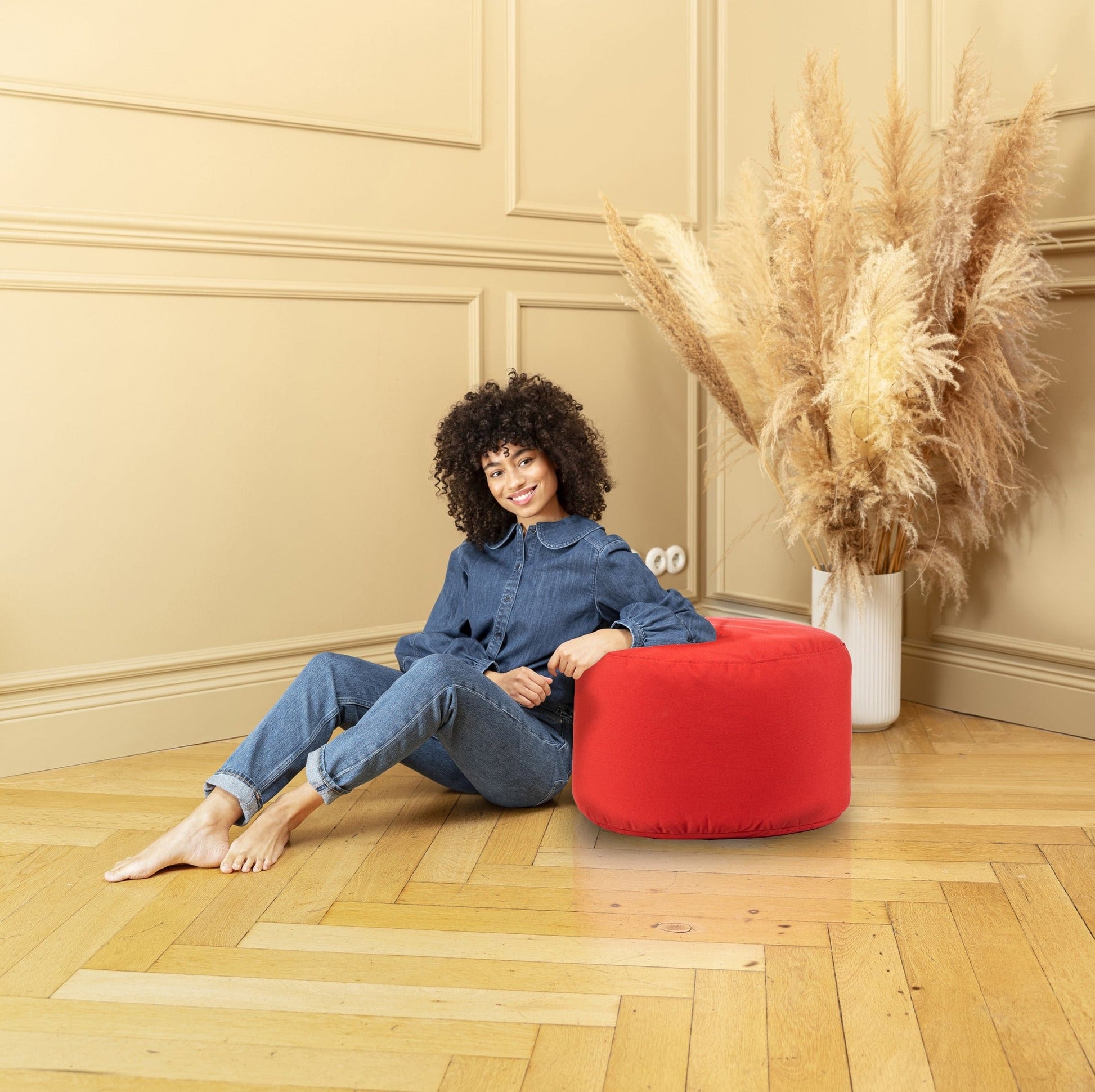 Woman sitting on a red ottoman in a room with beige walls and a vase of dried plants.