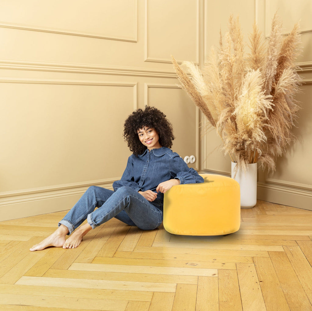 Woman sitting on a yellow ottoman in a room with beige walls and a vase of dried plants.