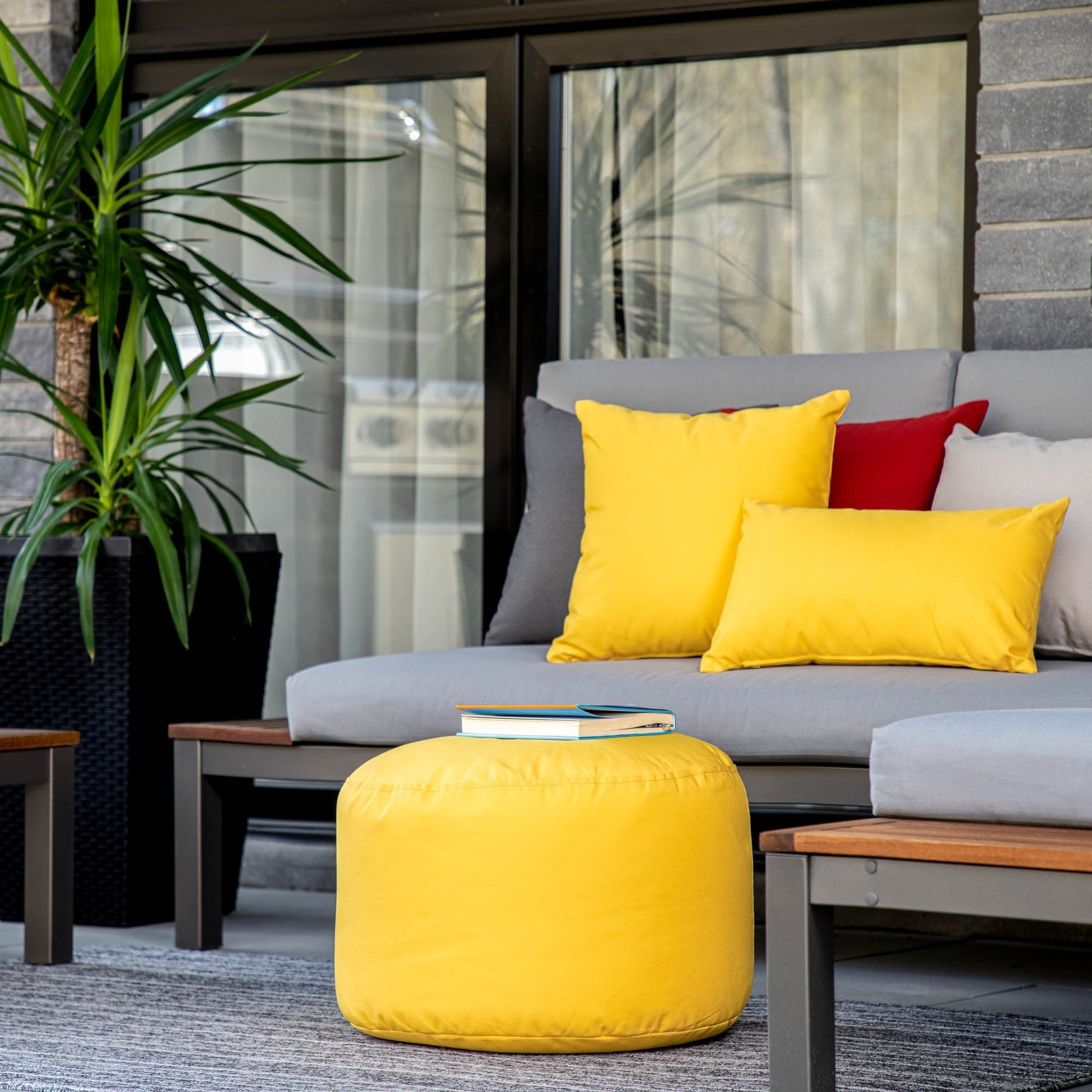 Outdoor seating area with a gray sofa, yellow pouf, and colorful cushions on a patio.