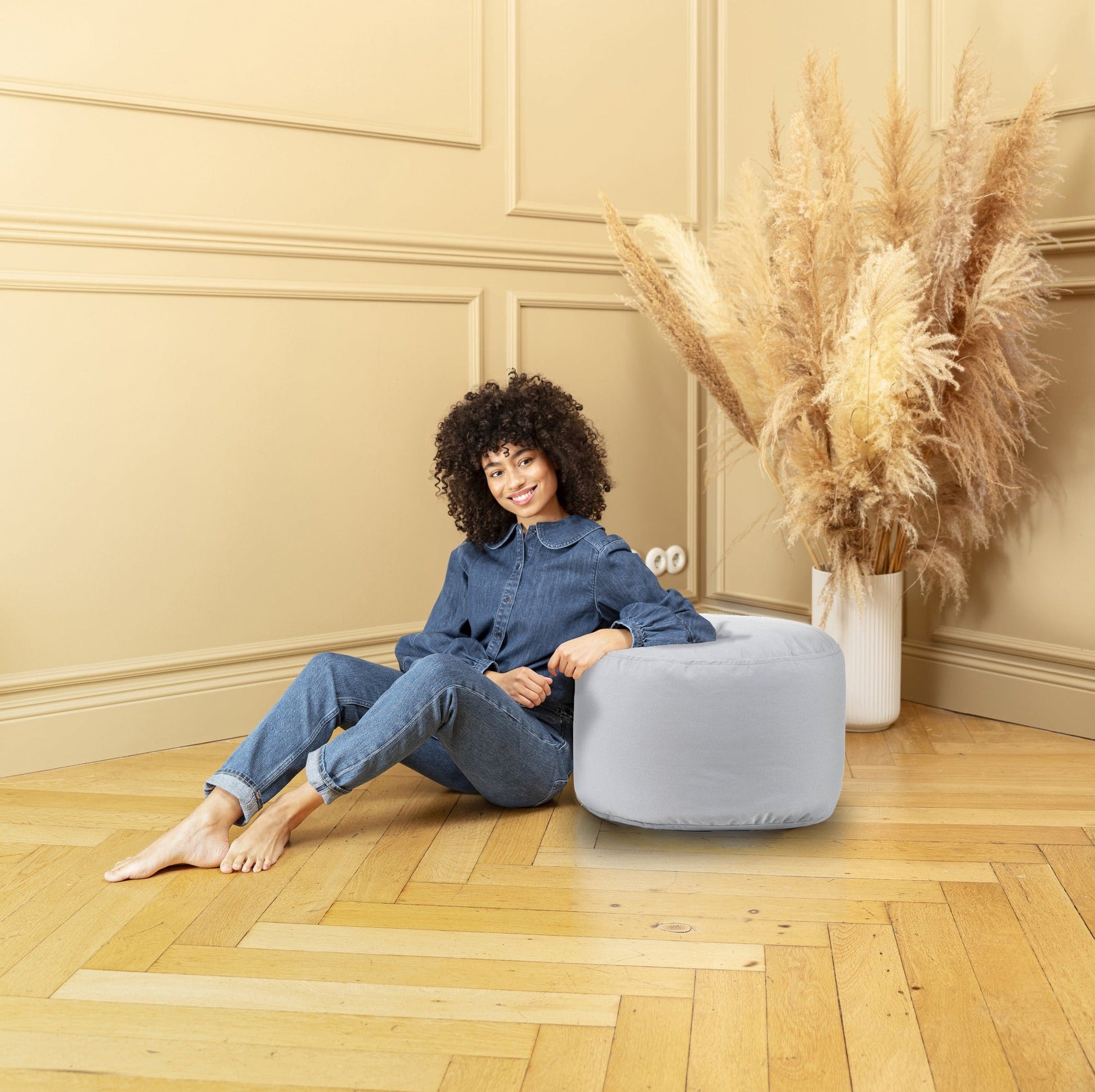 Woman sitting on a light gray pouf in a room with beige walls and wooden floor.
