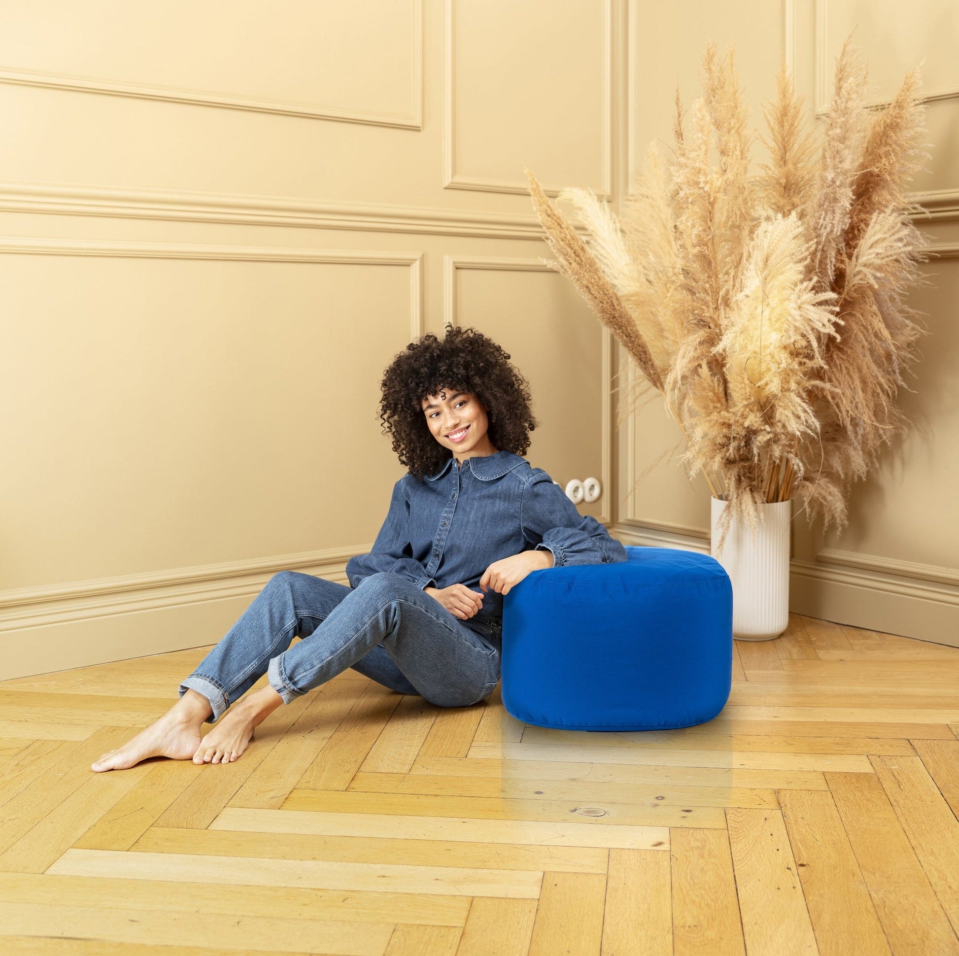 Woman sitting on a blue pouf in a room with beige walls and a vase of dried plants.