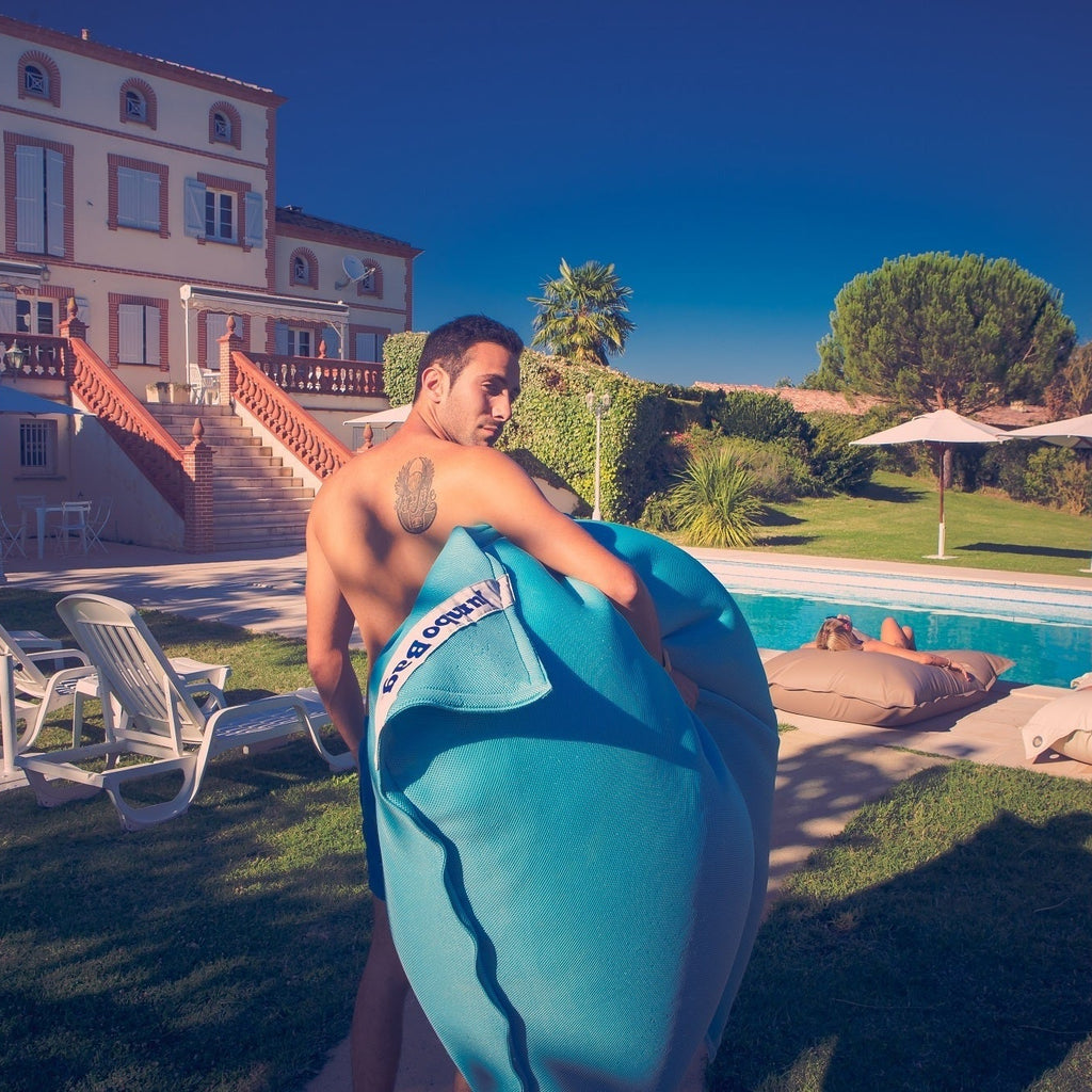 Man holding a blue inflatable raft by a pool with a large house in the background