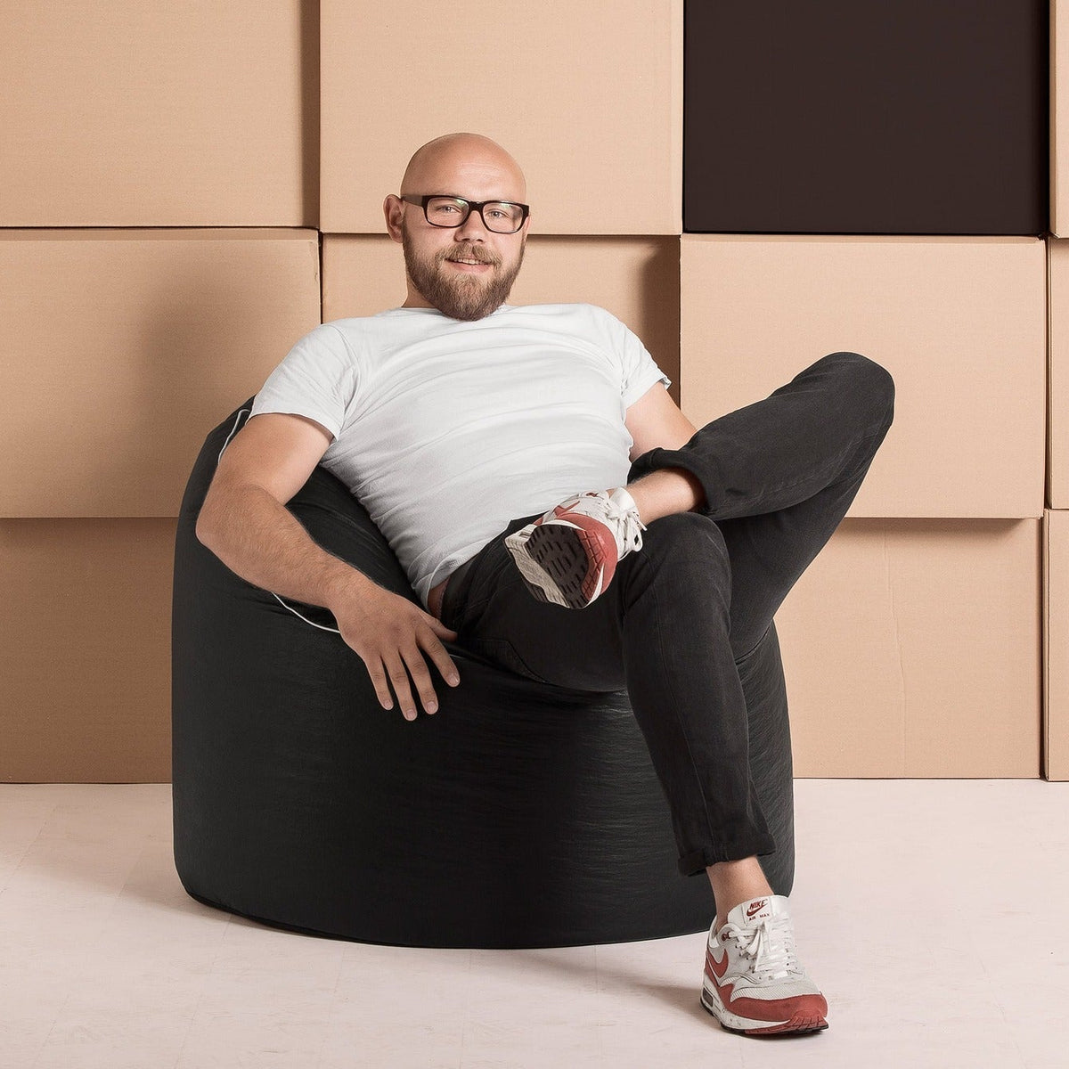 Man sitting on a black bean bag chair against a wall of cardboard boxes.